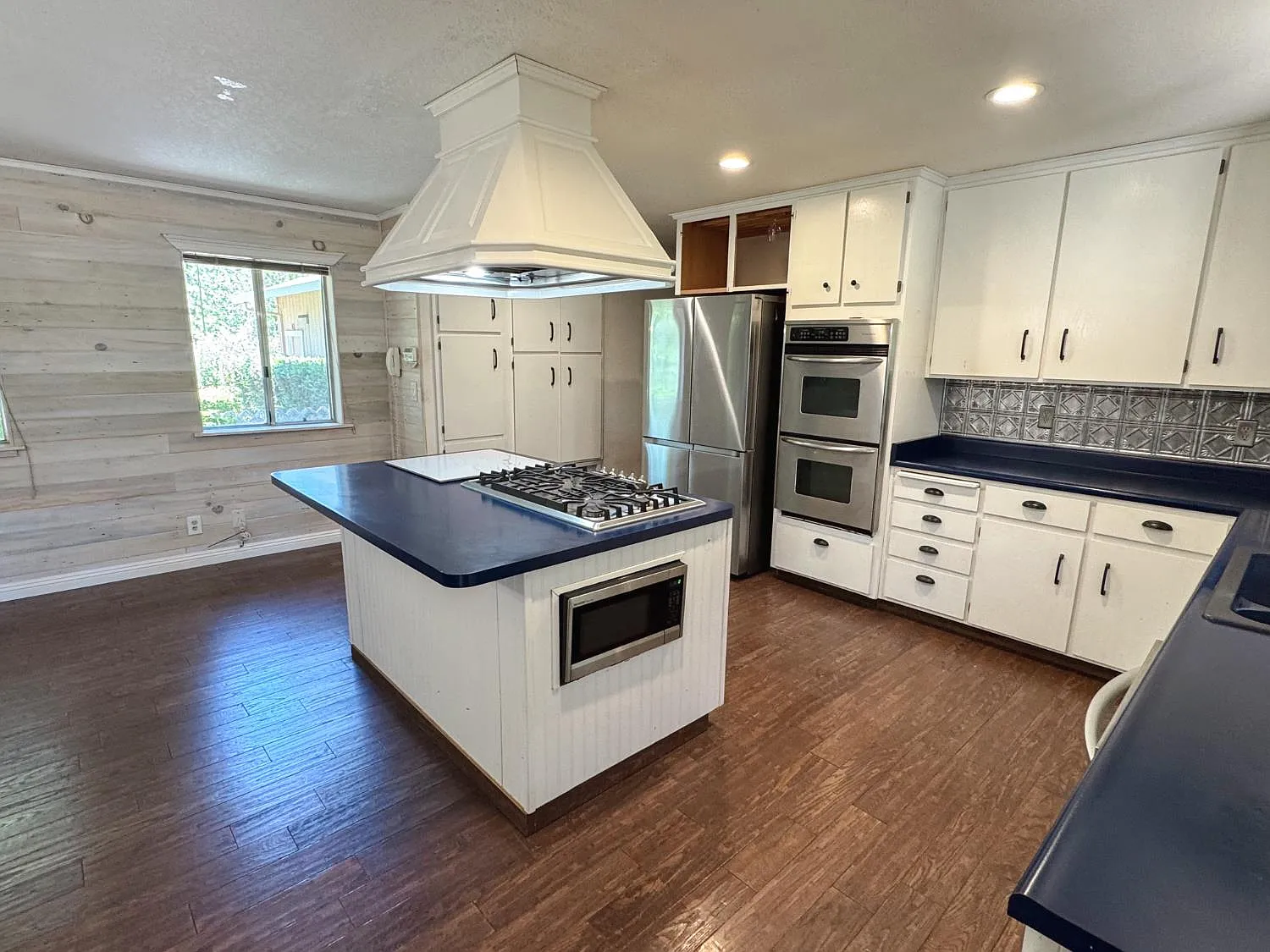 Kitchen with large central island, gas cooktop, vent hood, double wall ovens and white cabinetry